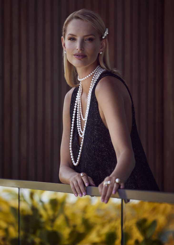 A white female blond model in a black strapless dress and a long string of pearls leans against a railing.