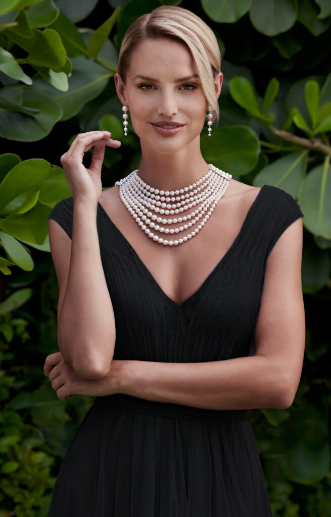 A white female model in a black dress wears a string of pearls and diamond, pearl earrings.