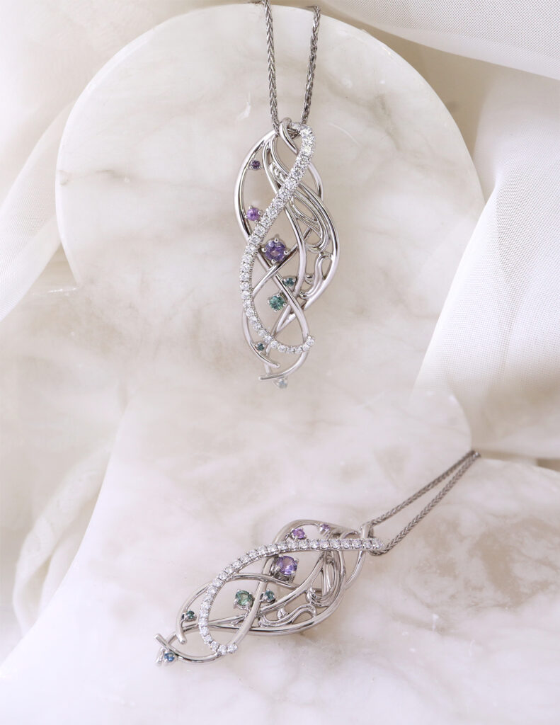 Two ornate pendants in white gold with diamonds and coloured sapphires on a white cloth background.