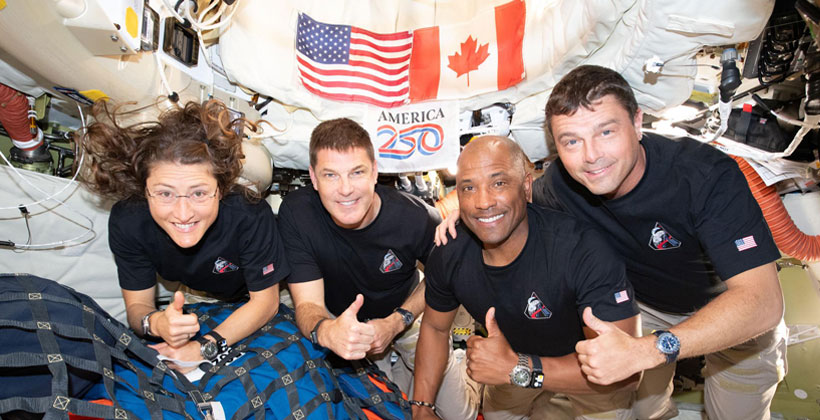 four people posing and hold thumbs up inside spacecraft