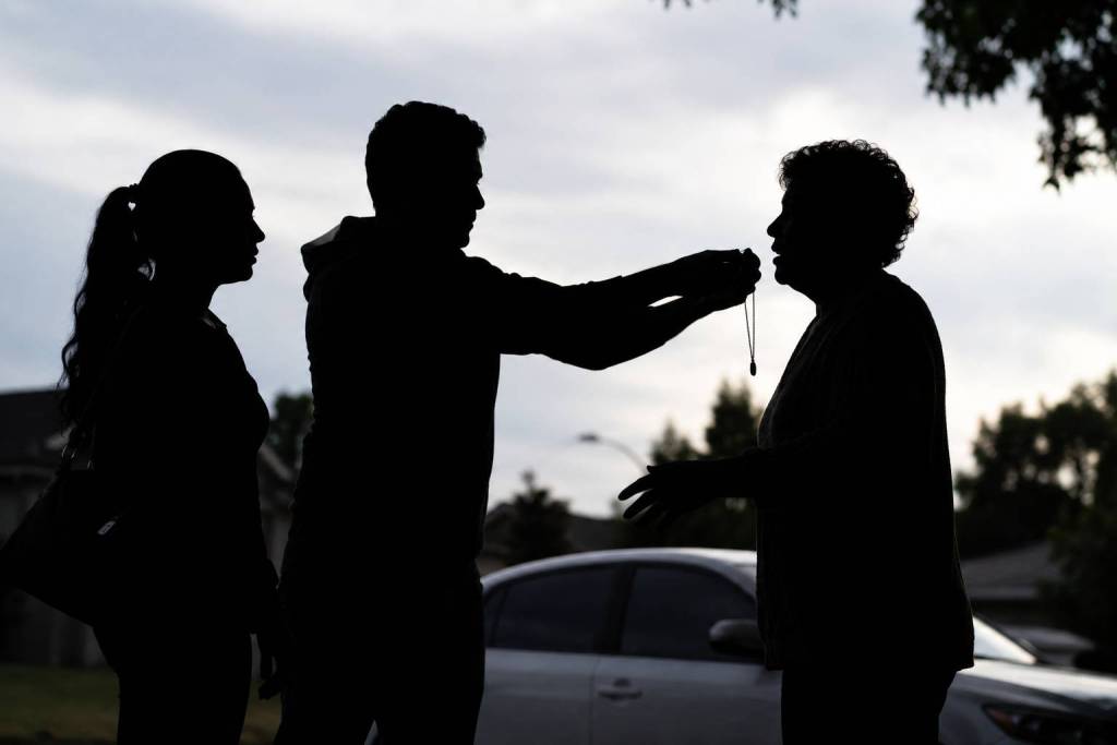 Three backlit silhouetted figures stand in a suburban front yard at dusk, with one person extending a necklace toward another while a third stands nearby, emphasizing a tense, staged exchange scene.