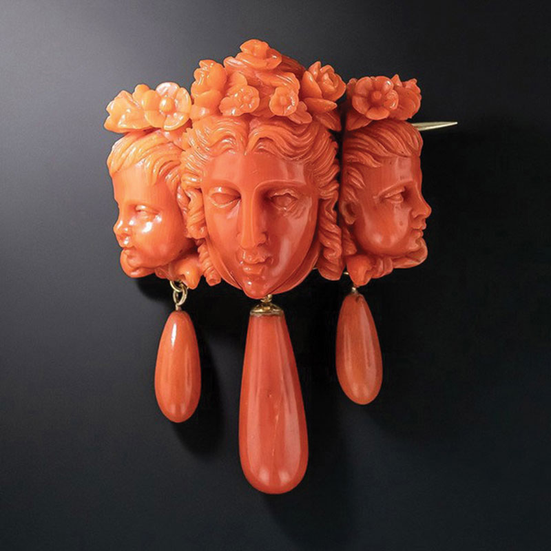 Carved coral brooch depicting three sculpted faces with floral hair details, each with a dangling coral teardrop.