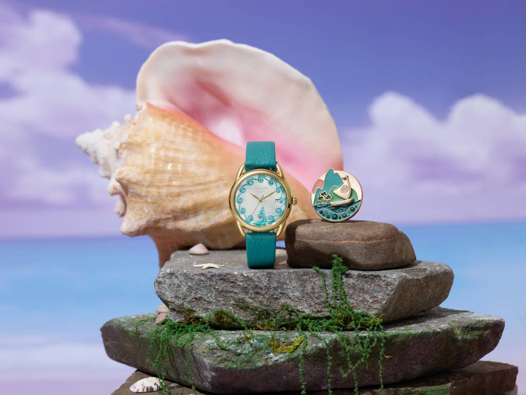 Product styling shot of the teal and gold Citizen watch displayed on stacked stones with moss, next to a round gold pendant charm depicting a green mountain, palm trees, and a sailboat with teal gemstone accents. A large pink-and-cream conch shell and small starfish sit in the background against a purple sky and blue ocean backdrop.