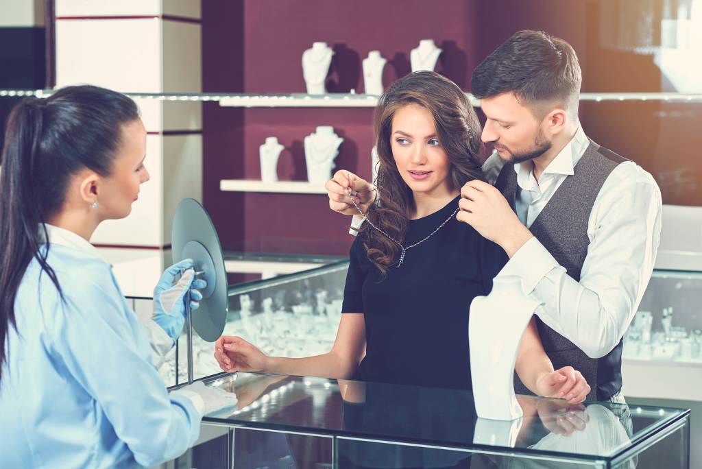 A stock image of a couple buying jewellery with the seller holding a mirror as they try on a necklace.