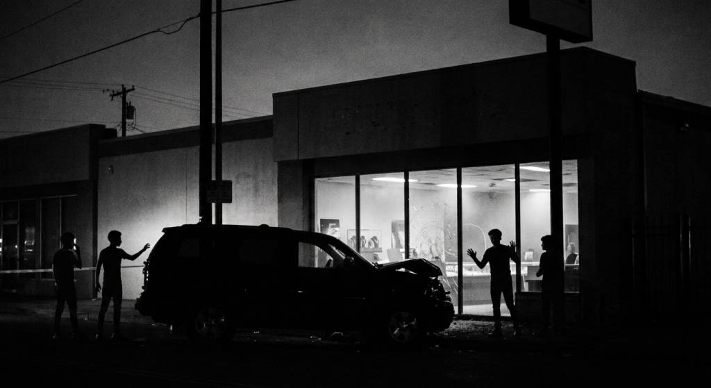 Black-and-white silhouette of young men standing outside a jewelry store at night next to an SUV crashed into a cracked glass storefront.
