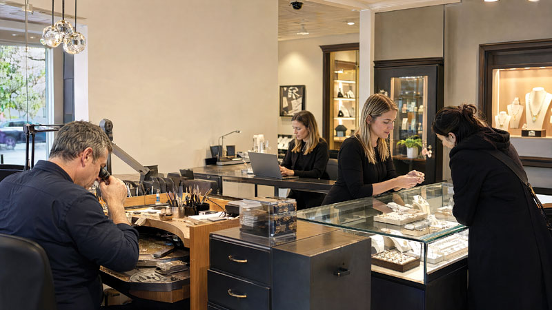 Inside a jewellery store, a bench jeweller works with a loupe at left while two staff members help a customer looking at rings in a glass display case.